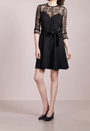 Woman wearing a knee-length black dress with lace 3/4 sleeves and a tied waist belt, paired with black high-heeled shoes.