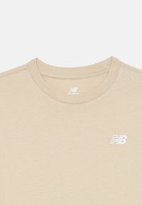 T-shirt de gola redonda bege da New Balance com o logótipo bordado "NB" em branco no peito e o nome da marca impresso dentro do colarinho.