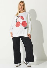 Överdimensionerad vit sweatshirt med en röd körsbärsgrafik och texten "Cherry Bomb". Kombinerad med vida svarta byxor och sportiga skor.