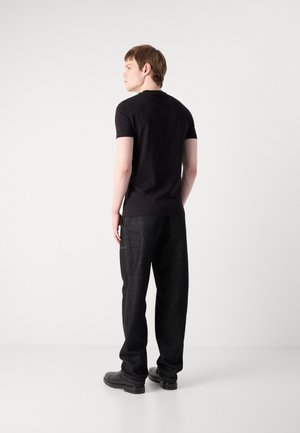 Jeune homme debout, face à gauche, portant un t-shirt noir à manches courtes, un pantalon ample foncé et des chaussures noires sur un fond clair uni.