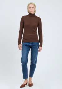Brauner Rollkragenpullover mit langen Ärmeln, kombiniert mit hellblauen Jeans und braunen spitz zulaufenden Schlüpfschuhen. Glatte Textur, taillierter Schnitt.