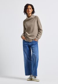 Beige gebreide trui met een brede col, geribbelde accenten en knopen; gecombineerd met losse blauwe jeans en lichtgekleurde sneakers.