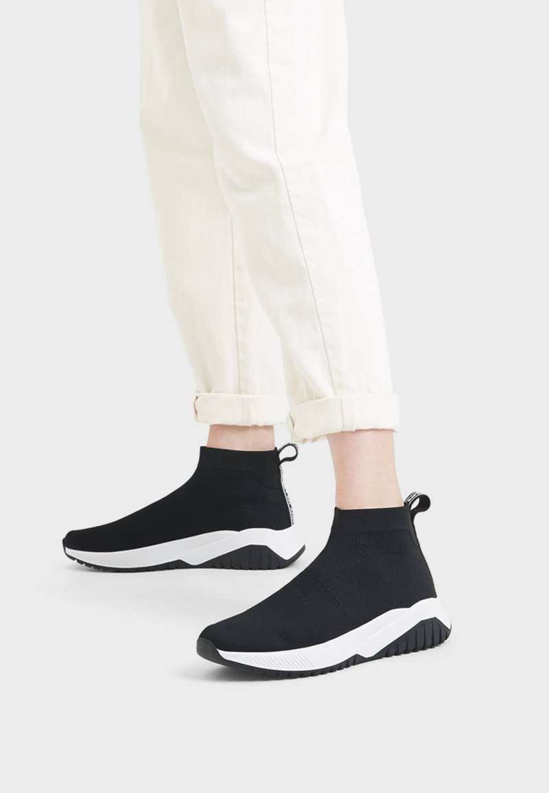 Sneakers alte nere in maglia con suola bianca, tomaia testurizzata e linguetta sul tallone, abbinate a pantaloni bianchi arrotolati.