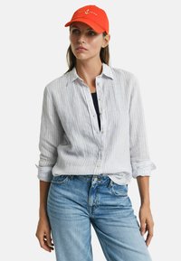 Lichtblauw gestreept linnen shirt met opgerolde mouwen, gecombineerd met vervagende blauwe denim jeans. Oranje honkbalpet met een ankerlogo.