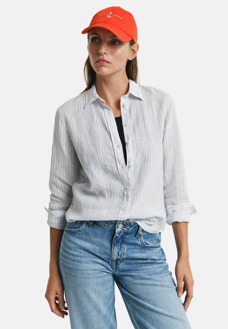 Camisa de lino a rayas azul claro con las mangas remangadas, combinada con unos vaqueros de denim azul descolorido. Gorra de béisbol naranja con un logo de ancla.
