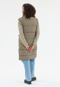 Olivgrüner, gepufferter Westen mit strukturiertem Design, knielang und mit Seitentaschen, getragen über einem hellgrauen Langarmshirt und blauen Jeans.