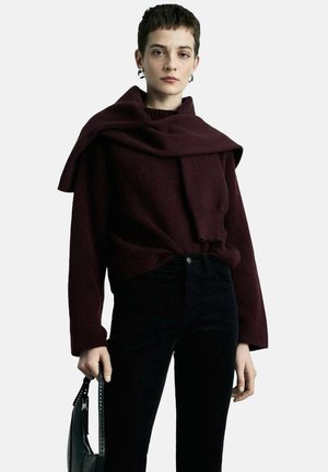 Burgundy knit sweater with a folded collar, paired with high-waisted black jeans. Model holds a black bag, showcasing textured materials.