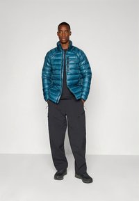 Tīrkizzaļš pūkains jaka ar horizontālu šūtu rakstu, ar rāvējslēdzēja kabatu uz krūtīm; kopā ar melnas krāsas kravas biksēm un atbilstošiem zābakiem uz vienkārša fona.