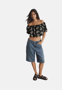 Zwarte off-shoulder crop top met bloemmotief, gecombineerd met lichtblauwe denim shorts en zwarte slide sandals. Soepele stof en relaxed fit.