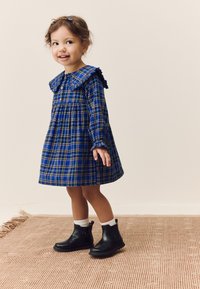 Robe à carreaux bleus avec col à volants, manches longues et devant boutonné. Bottines noires à fermeture éclair et chaussettes blanches aux chevilles.