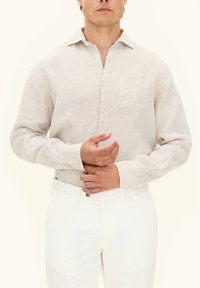 Camicia in lino beige con maniche lunghe, colletto a punta e taschino frontale; abbinata a pantaloni bianchi. Sottile texture e aspetto di tessuto naturale.
