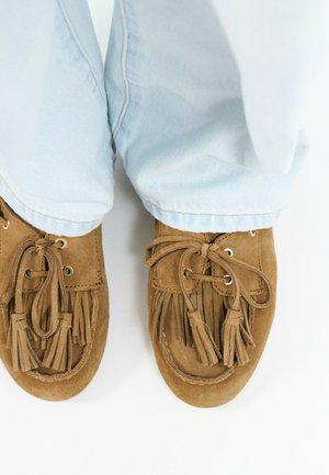 Mocasines de gamuza marrón con borlas, usados con jeans de mezclilla azul claro sobre un fondo blanco.