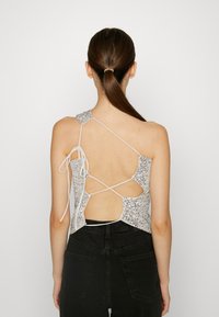 Silver sequin tank top with an open back, featuring white ties, asymmetrical design, and a fitted shape. Worn with high-waisted black pants.