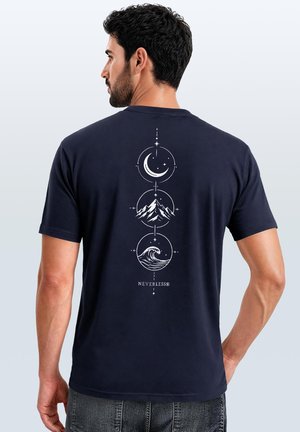 Mann trägt ein dunkelblaues Hemd mit weißen Symbolen von Mond, Berg und Welle, die vertikal am Rücken über dem Schriftzug "NEVERLESS®" angeordnet sind.