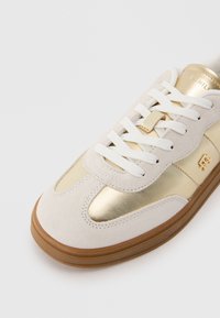 Sneaker din suede alb cu accente metalice aurii, având vârful rotunjit, șireturi plate albe și talpă din cauciuc.