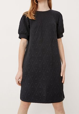 Femme portant une robe noire au genou avec des manches courtes bouffantes et un motif triangulaire texturé sur le tissu.