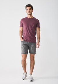 Burgunderfarget t-skjorte med korte ermer, kombinert med rullet opp grå shorts og hvite sportssko; modellen står med hendene i lommer mot en ensfarget bakgrunn.
