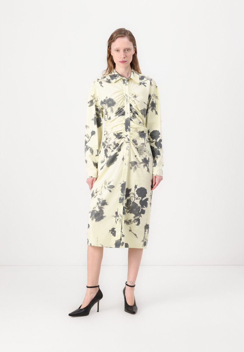 Long-sleeve, button-up dress in pale yellow with black floral print, featuring a ruched front and straight silhouette, paired with black heels.