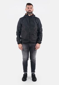 Giacca leggera nera con cappuccio, dotata di zipper frontale e due tasche laterali; indossata sopra jeans grigi e sneakers nere.