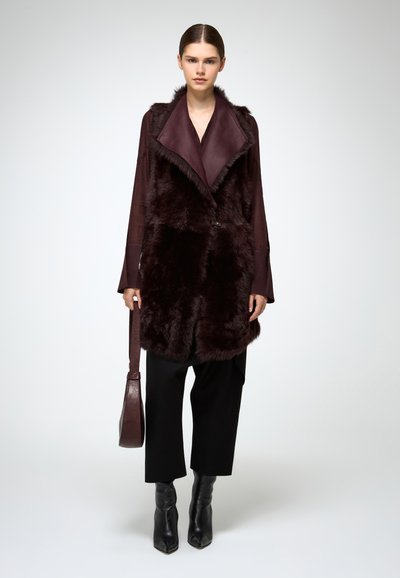 Donna che indossa un cappotto di pelliccia bordeaux scuro sopra una maglietta a maniche lunghe, pantaloni neri a vita alta, stivaletti neri, e tiene una borsa bordeaux abbinata su uno sfondo semplice.