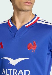 Modrý dres s červenými a bílými akcenty; obsahuje vyšívané logo kohouta, límec s kontrastními proužky a bílé logo sponzora.