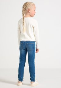 Dieťa s dlhými vrkôčikmi v blond farbe, oblečené v bielom svetri, modrých džínsach a bielych teniskách, stojí chrbtom oproti jednoduchému pozadiu.