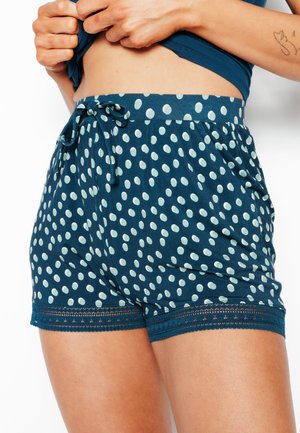 Vrouw draagt teal korte broek met witte stippen en kanten randje, met een zijzak en een klein strikje aan de tailleband.