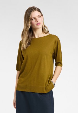 Kobieta o długich blond włosach, ubrana w musztardowo-zieloną bluzkę z krótkim rękawem i ciemną spódnicę, z jedną ręką w kieszeni na jednolitym jasnym tle.