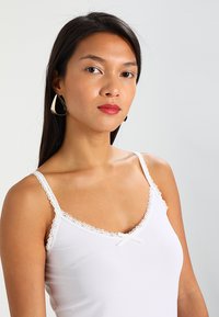 Camisola bianca con pizzo lungo il collo e le spalline, impreziosita da un piccolo fiocco al centro. Tessuto dalla texture liscia.