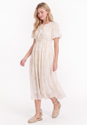 Blondynka ubrana w jasnobeżową midi sukienkę z bufiastymi rękawami i espadryle na płaskim obcasie z wiązaniami wokół kostek, stojąca na białym tle.