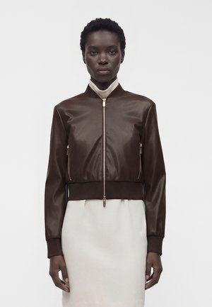 Woman with short curly hair wears a dark brown zip-up leather jacket over a high-neck white dress, standing against a plain background.