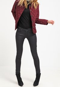 Bordeauxkleurig gewatteerd jack met knoopsluitingen, zwart top met lange mouwen, donkere skinny jeans en zwarte enkellaarsjes. Gladde texturen, aansluitend ontwerp.