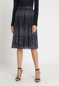 Jupe midi bleu marine en sequins avec un design plissé, dotée d'une taille ajustée. Associée à des sandales à talons hauts noires à lanières.