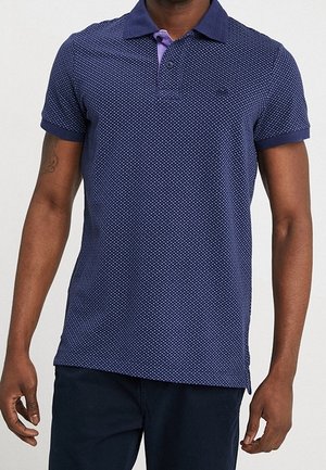 Polo azul marino con pequeños lunares blancos, mangas cortas, cuello morado contrastante y cierre con un solo botón en el cuello.