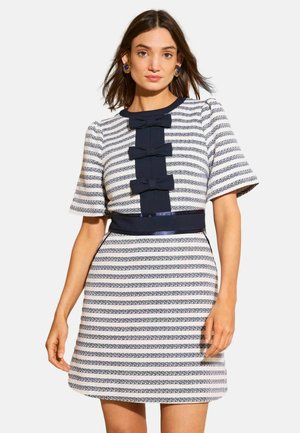 Femme portant une robe blanche à manches courtes avec des rayures horizontales bleu marine, des nœuds bleu marine à l'avant et une ceinture bleu marine, debout devant un fond uni.