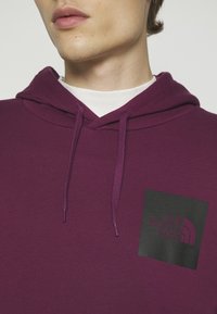 Personne portant un sweat à capuche violet The North Face avec cordons sur une chemise blanche, montrant uniquement la partie inférieure du visage et le cou.