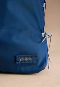 Námornícky modrá nylonová taška s hladkým povrchom, s zlatou nálepkou "Cath Kidston" a sivými viazacími akcentmi na boku.