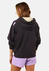 Haut à capuche noir avec un revêtement beige clair et une étiquette violette. Présente une coupe décontractée, une grande capuche et des poignets côtelés. Porté avec un short lavande.