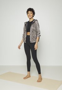 Leopárdmintás cipzáras dzseki, könnyű anyag, fekete crop top, fekete leggings, és mezítláb a bézs jógaszőnyegen, sima háttér előtt.