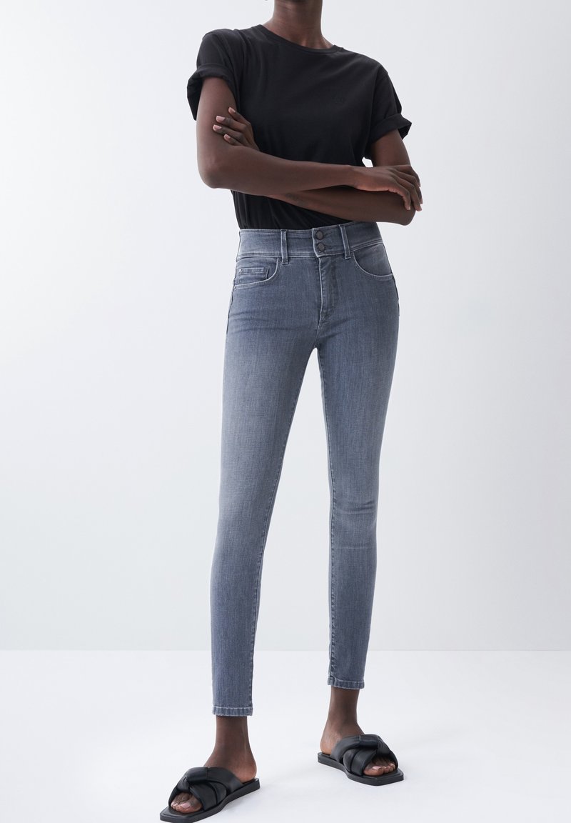 Jeans pitillo en un denim gris medio, diseño de talle alto, con dos bolsillos delanteros y costuras contrastantes. Se llevan con una camiseta negra y sandalias.