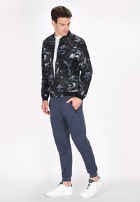 Zwarte bomberjack met blauw bloemenpatroon, gecombineerd met marineblauwe joggers, witte sneakers en een lichtgrijze shirt; fashionable sportieve kleding.
