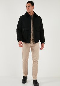 Sort bomberjakke med pelskrave, lynlåslukning foran, to forlommer, båret over en beige sweater og beige bukser, kompletteret med sorte sneakers.