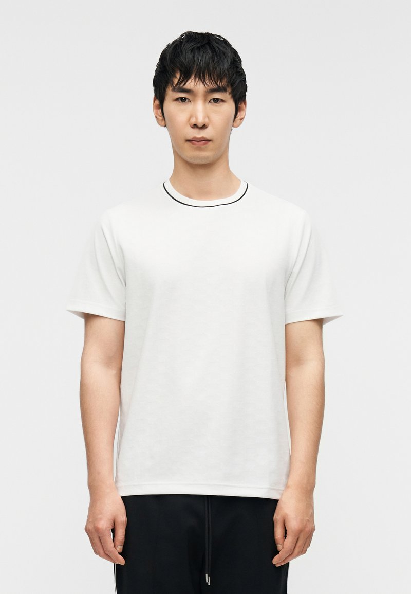 Λευκό T-shirt με στρογγυλή λαιμόκοψη που διαθέτει μαύρη λεπτομέρεια, κοντά μανίκια και διακριτικό υφασμάτινο μοτίβο, συνδυασμένο με σκουρόχρωμο παντελόνι.