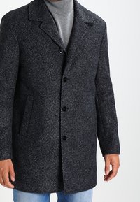 Abrigo de lana en diseño de espiga gris y negro con cuello de muesca, cierre de botón y bolsillos laterales, llevado sobre un suéter de cuello alto gris.