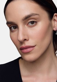 Zbliżenie portretu osoby o jasnobrązowej karnacji, noszącej małe srebrne kolczyki w kształcie kółek, z gładką skórą i naturalnym makijażem.