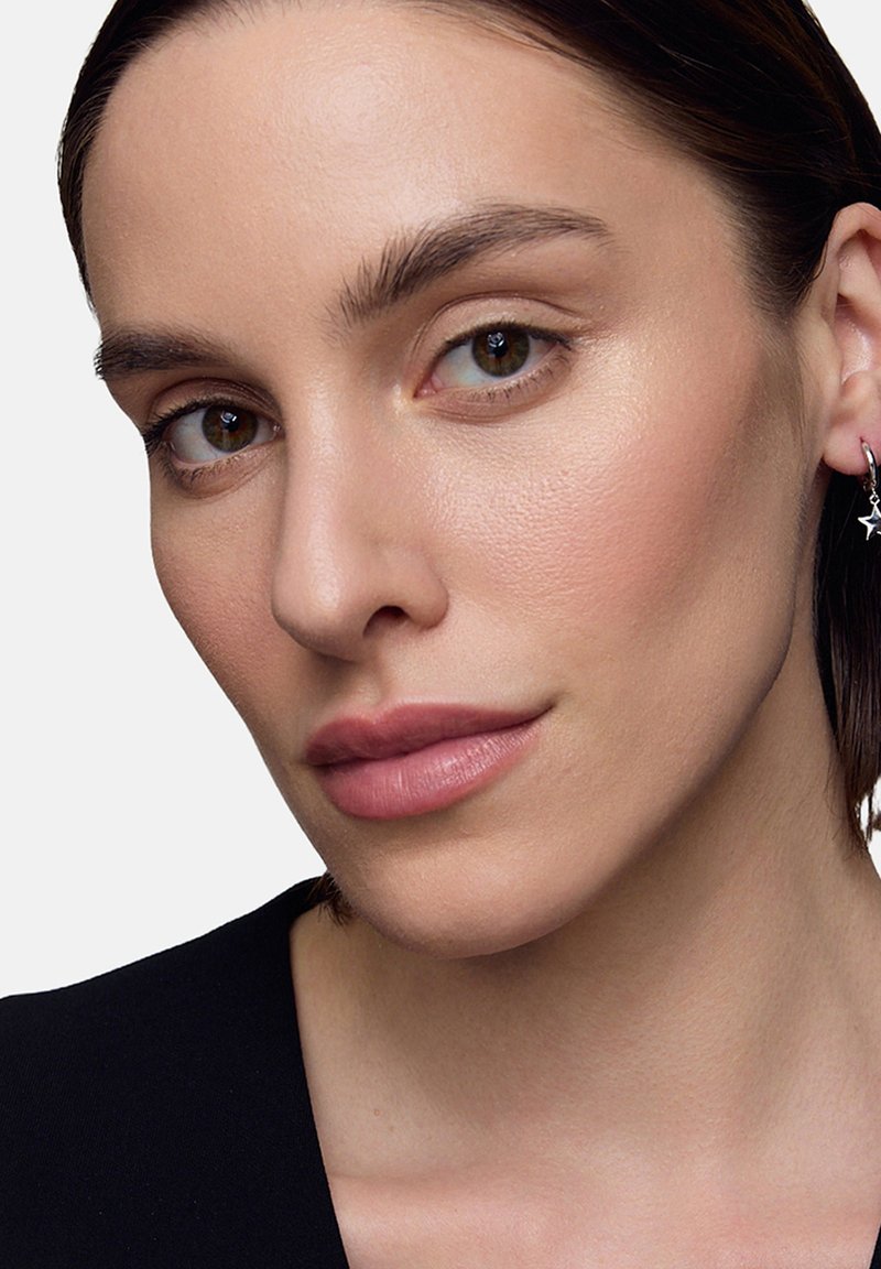 Zbliżenie portretu osoby o jasnobrązowej karnacji, noszącej małe srebrne kolczyki w kształcie kółek, z gładką skórą i naturalnym makijażem.