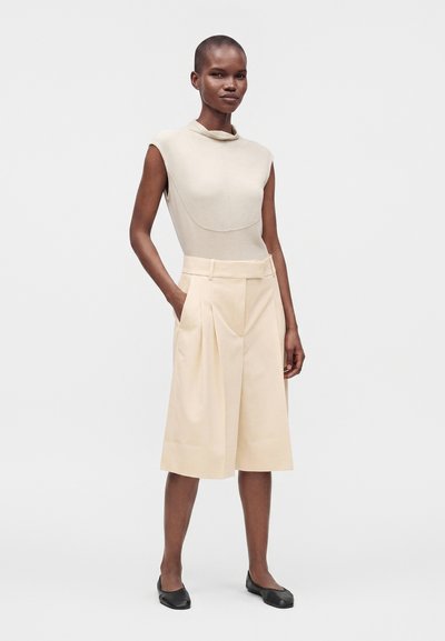 Femme mince aux cheveux courts portant un haut beige sans manches, un short large beige jusqu'aux genoux avec des poches, et des chaussures plates noires sur un fond uni.