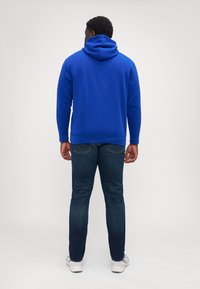Hanorac albastru cu buzunar tip kangur, glugă cu șnur și manșete ribbate. Purtați cu blugi închiși la culoare și pantofi sport de culoare deschisă. Vedere din spate.