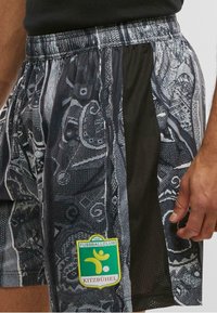 Sorte og grå mønstrede shorts med et mesh-panel og et grønt og gult logo fra Fussballclub Kitzbühel på venstre ben.