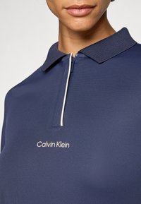 Námorníckom modrá polo košeľa s textúrovaným golierom, s polovičným zipsom a svetloružovým akcentom, s vyšitým logom "Calvin Klein" v bielej farbe.
